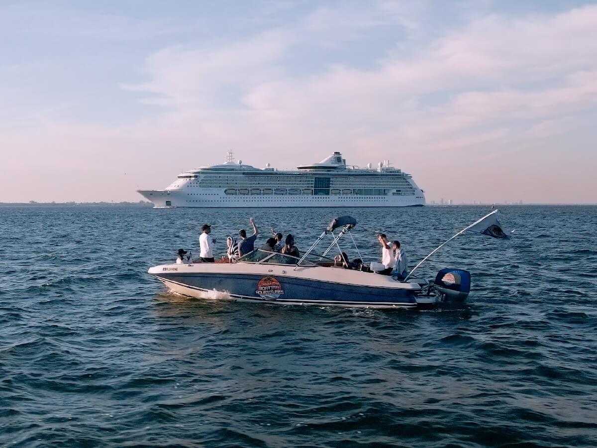 The charter broadside on open bay water with guests waving and a cruise ship on the horizon at dusk.
